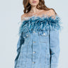A woman poses in the CHIC AF DENIM MINI DRESS WITH FEATHERS. Her long brown hair cascades down against a plain background, showcasing her effortless style with an off-the-shoulder design and blue feathers along the neckline.